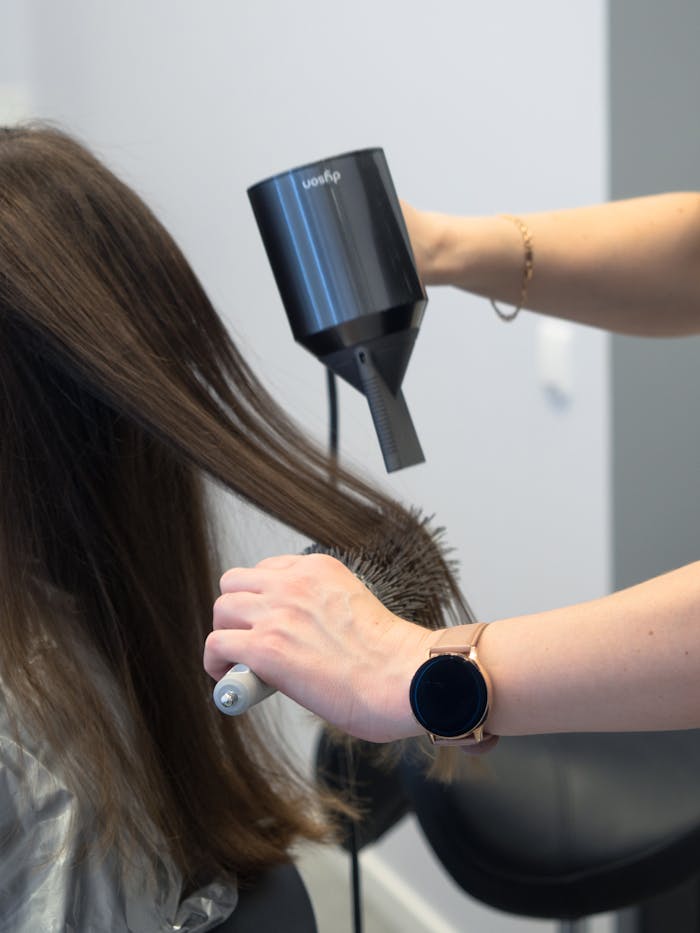 hairdresser-styling-hair-with-a-hairdryer-13714794 Close-up of a hairdresser using a hairdryer and brush for styling in a salon.